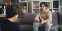 A teenager is sitting on a couch in a comfortable therapeutic space talking with a counsellor who is holding a clipboard.